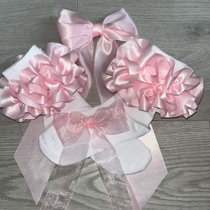 Puede incluir: Un par de calcetines blancos con grandes volantes de satén rosa claro superpuestos en los puños. Un lazo de organza rosa claro transparente está sujeto al centro de los calcetines, y un lazo de satén rosa claro más grande se sitúa encima.