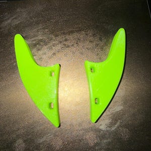 May include: Two neon green, 3D-printed objects, possibly components for a larger device, are shown on a dark surface. Each piece has a curved, wing-like shape with three small holes. The objects are brightly coloured and appear to be made of plastic.