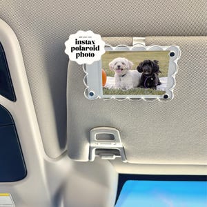May include: A clear plastic car visor photo holder with a scalloped edge, holding a photo of two dogs. The photo holder has a metal clip and the text "add your own instax polaroid photo".