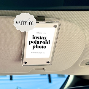 May include: A clear acrylic Instax Polaroid photo holder with the text "add your own instax polaroid photo" and the brand name "made by matty & co." The holder is attached to a black clip.