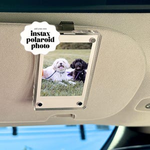 May include: Clear acrylic Instax Polaroid photo holder with a black clip, hanging from a car visor. The photo shows two dogs on grass. The holder has the text "add your own instax polaroid photo" in a white, starburst-shaped design.