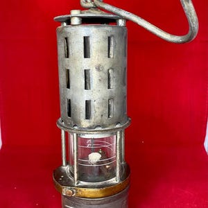 Può includere: Lampada da minatore vintage in metallo con corpo cilindrico e manico curvo. La lampada presenta un involucro di vetro e una fascia color ottone. Il corpo ha ritagli rettangolari. Su sfondo rosso.