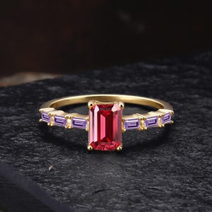 May include: A gold ring featuring a large, rectangular, red gemstone as the centerpiece. The ring is further adorned with smaller, rectangular, purple gemstones on either side of the main stone, creating a striking contrast against the gold band.