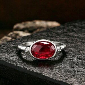 Puede incluir: Un anillo de plata con una gran piedra preciosa de color rojo intenso y corte ovalado en el centro. La banda está decorada con dos pequeñas piedras claras de corte redondo a cada lado del engaste central, enmarcado por un borde metálico texturizado.