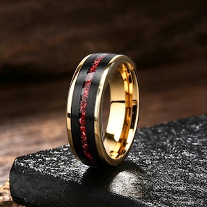 Pode incluir: Um anel com interior e bordas douradas polidas, apresentando uma faixa preta larga com acabamento escovado e uma incrustação central de fragmentos vermelhos brilhantes. O design combina superfícies de ouro metálico e preto fosco com um centro.