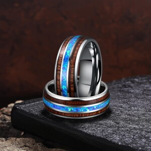Puede incluir: Dos anillos de metal con una incrustación central de ópalo azul vibrante flanqueada por madera de color marrón oscuro. Los anillos tienen un acabado plateado pulido en los bordes, creando una superficie lisa y reflectante que contrasta.