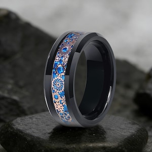 May include: A black ring with a blue and bronze gear design. The ring has a polished black band with a detailed pattern of gears and floral elements in blue and bronze. The ring is displayed on a dark, textured surface.