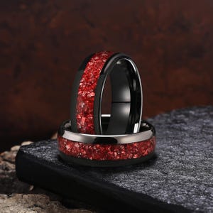 Può includere: Due anelli neri con un intarsio centrale di materiale rosso frantumato. Un anello è posizionato verticalmente mentre l'altro poggia orizzontalmente sotto di esso. Le fasce presentano bordi lisci e lucidi che contrastano con l'intarsio rosso testurizzato.