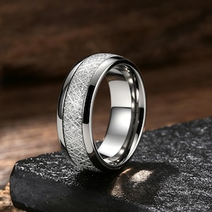 Può includere: Un anello in metallo color argento caratterizzato da un'ampia fascia centrale testurizzata con una finitura scintillante a tratteggio incrociato. L'anello presenta bordi lisci e lucidi su entrambi i lati, creando una superficie riflettente che contrasta con la texture opaca e granulosa dell'inserto centrale.