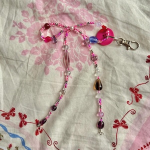 May include: A beaded keychain with a pink bow design. The keychain features a variety of beads in shades of pink, purple, and clear, with a pink button and silver clasp. The background is a pink floral pattern.