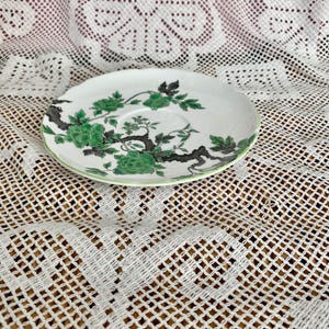 May include: A white saucer with a green floral pattern and a green edge. The floral design features green flowers and leaves with black stems. The saucer is on a white lace tablecloth.
