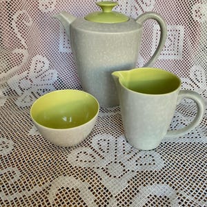 May include: A light grey speckled ceramic tea set with lime green detailing. The set comprises a teapot, a creamer jug, and a small bowl. The teapot has a green lid and a curved handle. The creamer and bowl have green interiors.