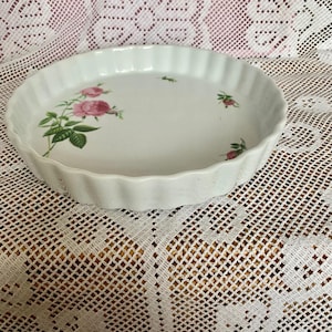May include: A white ceramic pie dish with a fluted edge. The dish features pink rose and green leaf designs. The round dish is approximately 23 cm in diameter.