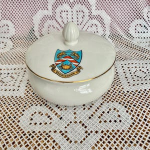 May include: A white porcelain lidded sugar bowl featuring a gold rim and a central crest with two lobsters and a crab above a banner reading SHERINGHAM. The lid has a small, pointed finial handle, and the bowl has a smooth, rounded body shape.