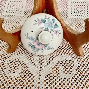 May include: A white ceramic lid decorated with a floral pattern in shades of blue and pink, with green foliage. The lid has a small knob and a small hole. Likely a teapot lid or similar.