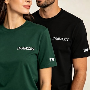Può includere: Una t-shirt verde scuro e una t-shirt nera, entrambe con il testo bianco I.V.MMXXIV sul petto. La maglietta verde presenta il testo bianco T seguito da un simbolo di cuore sulla manica, mentre quella nera ha D seguito da un cuore.