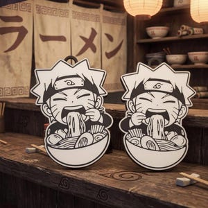 May include: Two black and white anime-style character ornaments. Each ornament depicts a character eating ramen from a bowl. The background includes a wooden surface, chopsticks, and Japanese text.