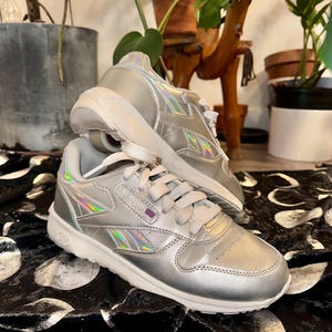 May include: Silver athletic shoes with iridescent accents and white soles. The shoes have a classic design with laces and the Reebok logo. The shoes are on a black and white patterned surface.
