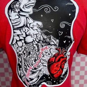 May include: Red t-shirt featuring a black and white graphic design on the back. The design includes a stylized human heart, swirling lines, and the text "Thousand Faces One Heart". The shirt's design is outlined in white.