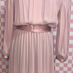Può includere: Un abito plissettato rosa chiaro con maniche lunghe e una cintura in raso abbinata. L'abito è esposto su un manichino nero su uno sfondo a scacchi rosa e bianco. L'abito ha una chiusura con bottoni sulla cintura.