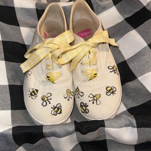 Puede incluir: Un par de zapatillas de lona blanquecinas decoradas con ilustraciones de abejas negras y amarillas. Los zapatos tienen cordones de cinta de cuadros amarillos y detalles en forma de abeja. Sobre un fondo a cuadros blanco y negro.