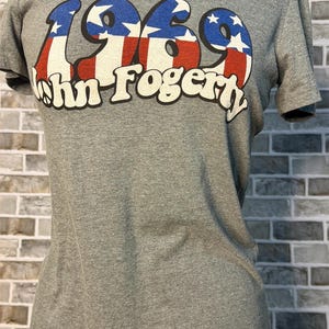 May include: A heather gray short-sleeve t-shirt featuring the text 1969 in a red, white, and blue star-spangled pattern. Below the year, the text John Fogerty is printed in white with a black outline, including a peace sign in the letter O.