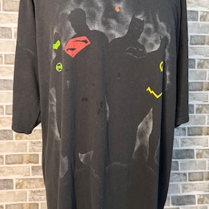 Puede incluir: Una camiseta negra de manga corta con un gráfico descolorido estilo pintura en aerosol que muestra siluetas de superhéroes. El diseño incluye un logotipo rojo de Superman, un emblema verde de Green Lantern y un rayo amarillo de Flash.