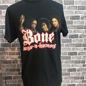 May include: Black t-shirt featuring a graphic of four people and the text "Bone thugs-n-harmony" in red and white. The shirt is displayed on a mannequin against a brick wall background.