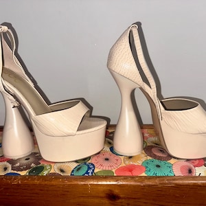 May include: A pair of beige platform sandals with a unique heel design. The sandals feature a textured upper, open toe, and ankle strap. The platform sole and sculptural heel add a bold, modern touch to these women's shoes.