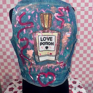 May include: A denim vest with a sequined "Love Potion" design. The back of the vest features a large perfume bottle with the words "Love Potion" in a square label. Pink paint splatters and a heart design complete the look.