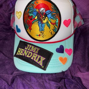 Puede incluir: Una gorra de camionero blanca y azul claro con un parche bordado circular de una figura con una guitarra caminando entre dos osos. La gorra incluye un parche rectangular negro con el texto JIMI HENDRIX en verde y morado, además de varios pequeños parches coloridos en forma de corazón.