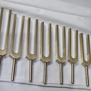 May include: A collection of ten gold-coloured tuning forks displayed on a white cloth. Each fork features a U-shaped top and a long, slender handle. The forks vary in size, indicating a range of musical notes.