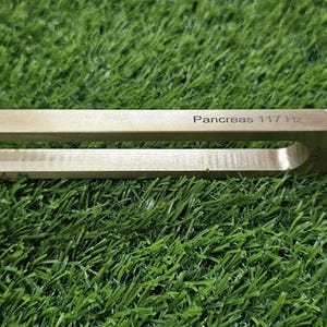 Peut inclure: Un diapason en métal doré doté de deux longues branches et d'un manche fin. Le côté du diapason porte l'inscription "Pancreas 117 Hz" gravée dans une police sombre et claire. L'objet présente une finition métallique lisse et une base en U.