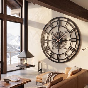 Puede incluir: Un gran reloj de pared circular de metal negro con un diseño de rosa de los vientos en el centro. La esfera muestra números romanos en el borde exterior y los puntos cardinales N, NE, E, SE, S, SW, W y NW. Tiene manecillas finas de color claro.