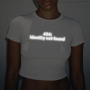 Op de afbeelding: Witte crop top met de tekst "404: identity not found" in een reflecterend, gloeiend lettertype. De top heeft korte mouwen en een ronde hals. De foto is gemaakt in een zwak verlichte omgeving.