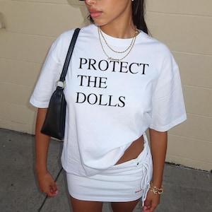 Peut inclure: Ensemble t-shirt et jupe blancs. Le t-shirt porte l'inscription noire "PROTECT THE DOLLS". La jupe a un détail de cordon de serrage sur le côté. Un sac à main noir est porté à l'épaule.