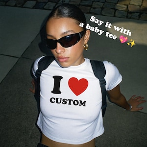Può includere: Baby tee bianca con la scritta "I CUSTOM" in nero e un cuore rosso. La maglietta ha anche la scritta "Say it with a baby tee" in bianco. La persona indossa occhiali da sole neri e orecchini a cerchio dorati.
