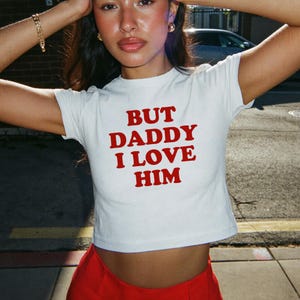 Op de afbeelding: Witte crop top met de tekst "BUT DADDY I LOVE HIM" in het rood. De top heeft korte mouwen en een ronde hals. Het model draagt een rode broek.