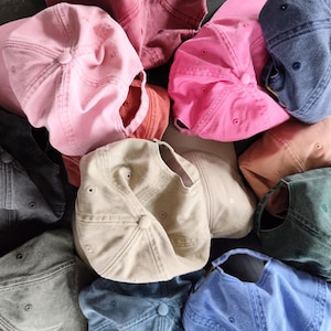 Puede incluir: Una colección de gorras de béisbol en varios colores, incluyendo rosa, azul, gris y beige. Las gorras están hechas de una tela suave y tienen un diseño clásico de seis paneles con una visera curva. Las gorras están apiladas.