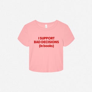 May include: A light pink short-sleeved crop top featuring the text "I SUPPORT BAD DECISIONS (in books)" printed in bold red capital letters across the center of the chest. The garment has a round neckline and a straight hemline.
