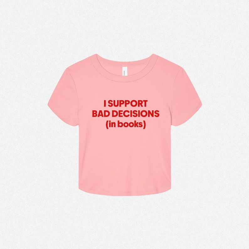 May include: A light pink short-sleeved crop top featuring the text "I SUPPORT BAD DECISIONS (in books)" printed in bold red capital letters across the center of the chest. The garment has a round neckline and a straight hemline.