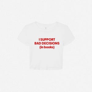 May include: A white short-sleeved crop top featuring the red text I SUPPORT BAD DECISIONS (in books) printed in a bold, sans-serif font across the center of the chest. The garment has a crew neckline and a cropped hemline.
