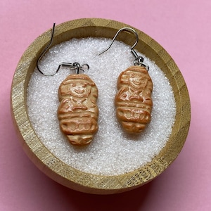 Peut inclure: Une paire de boucles d'oreilles de couleur beige avec un motif de figurine sculptée. Les boucles d'oreilles sont présentées dans un petit bol en bois rempli de granules blancs, sur fond rose. Les boucles ont des fermoirs en forme de crochet argentés.