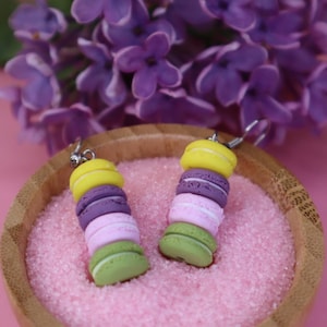 Può includere: Un paio di orecchini con macarons impilati in tonalità di giallo, viola, rosa e verde. Gli orecchini sono esposti su una superficie rosa con cristalli di zucchero.
