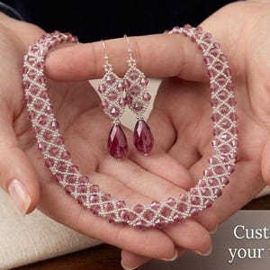 Puede incluir: Un collar y pendientes a juego hechos de pequeños cristales malva facetados en un patrón de rombos, con cristales en forma de lágrima en la base de cada pendiente. El texto dice: Customise it with your favorite color.