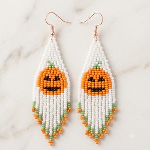 Può includere: Un paio di orecchini lunghi e pendenti realizzati con piccole perline. Il design presenta uno sfondo bianco con un volto di zucca arancione al centro, accentuato da perline verdi e arancioni sul bordo inferiore, appesi a ganci dorati.