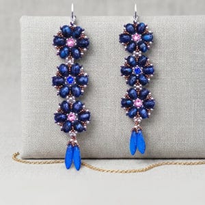 May include: A pair of long drop earrings featuring three vertically stacked flower motifs crafted from dark blue beads. Each flower center contains a small pink or blue crystal. The earrings terminate in two dangling blue petal-shaped beads.