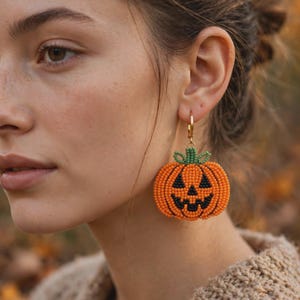 May include: A beaded earring shaped like a jack-o'-lantern pumpkin. The pumpkin is crafted from orange beads with a black beaded face featuring triangular eyes and a jagged mouth. A small green beaded stem sits on top, attached to a gold-tone hoop.