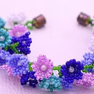 May include: A beaded bracelet featuring a floral design made of small glass beads in shades of blue, purple, turquoise, and white. The flowers are connected by green beaded stems, and the bracelet includes two metallic cylindrical end clasps.