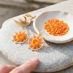 May include: A pair of sun-shaped dangle earrings featuring a central orange bead surrounded by radiating yellow and orange seed beads on a silver-toned circular frame, accompanied by a small bowl filled with matching loose orange and yellow beads.